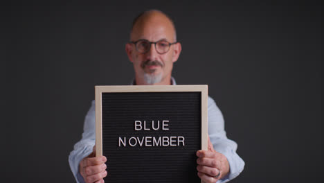 Studio-Portrait-Of-Mature-Man-Holding-Up-Sign-Reading-Blue-November-Promoting-Awareness-Of-Men's-Health-And-Cancer